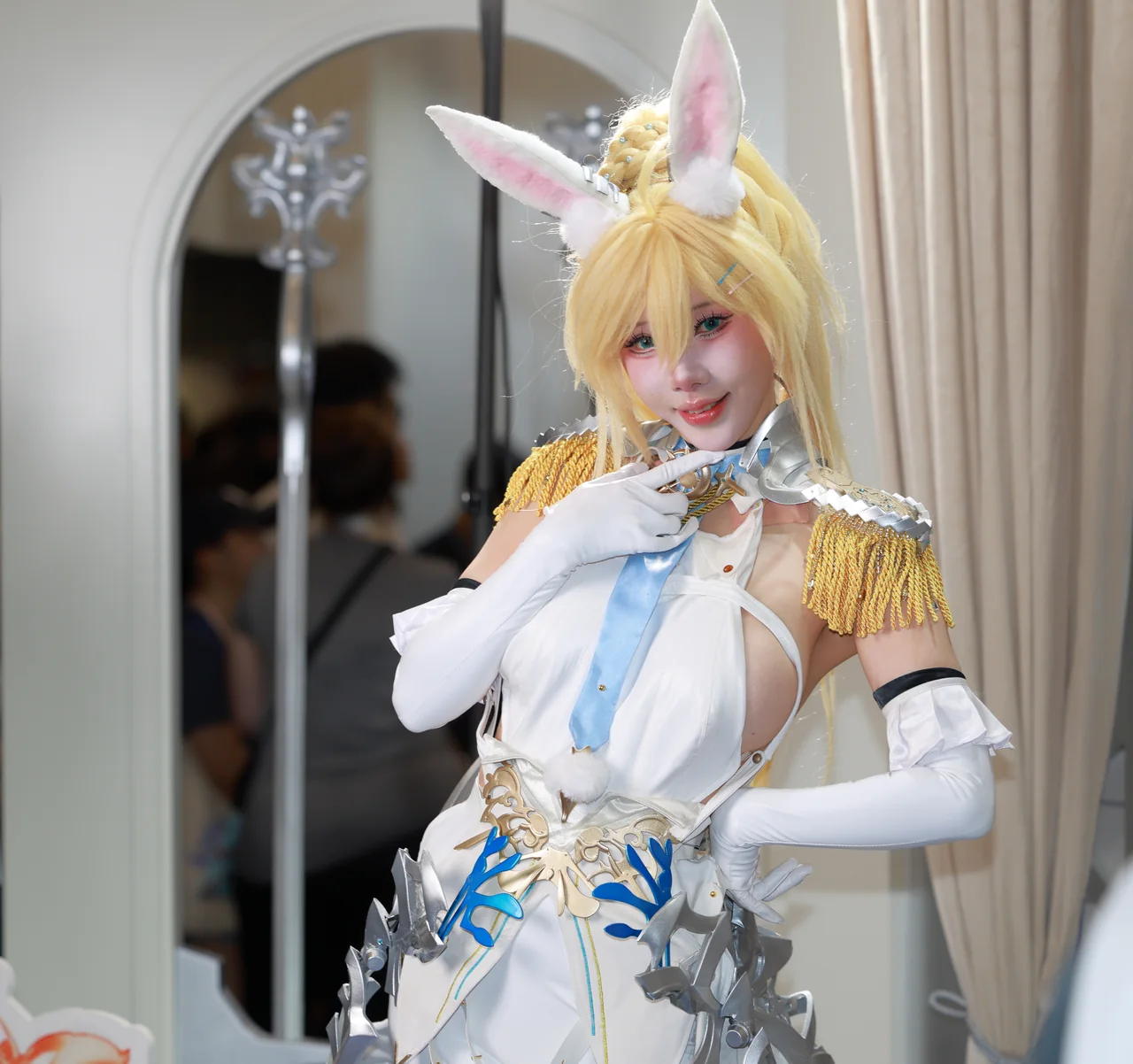 A blonde cosplayer in a white and gold costume posing beside a curtain