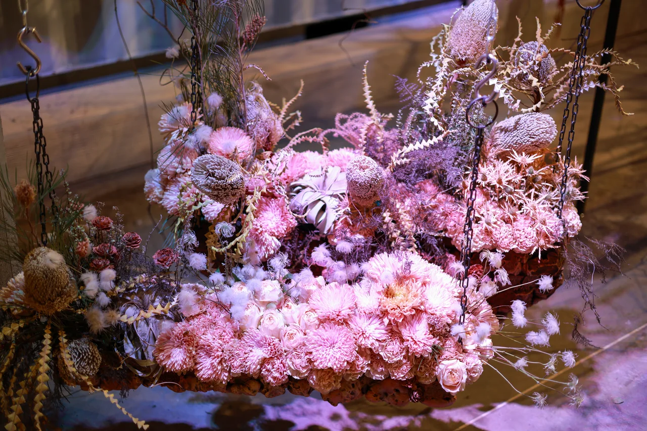 A suspended floral arrangement of pink and purple blooms, textured seed heads and delicate branches under violet light