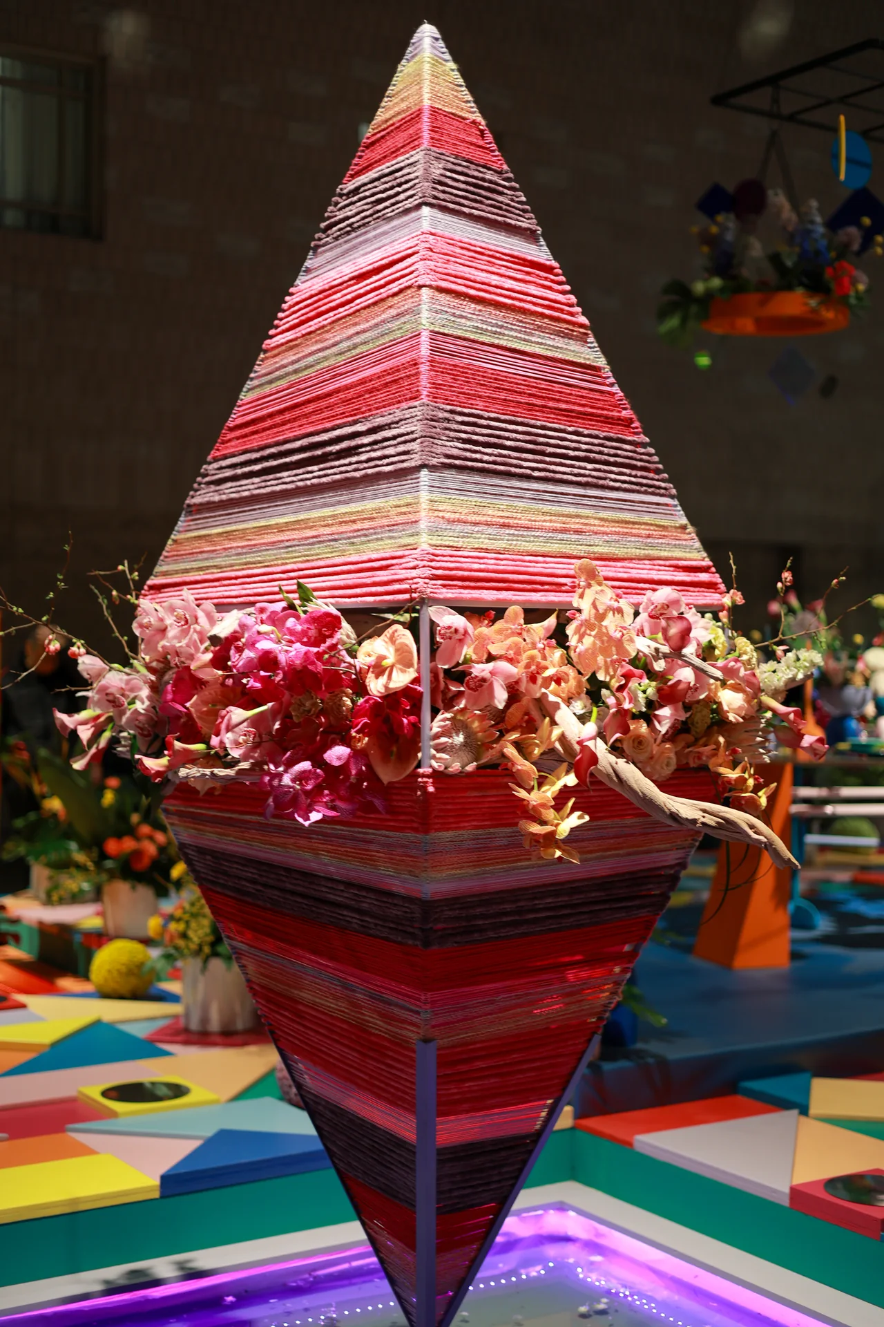 A tall diamond-shaped sculpture wrapped in colorful bands with flowers around its center