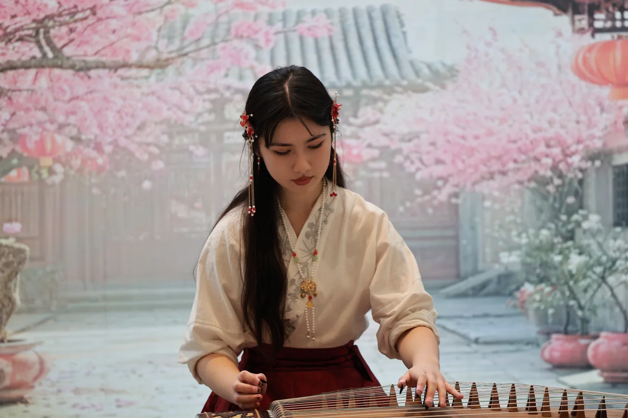 A musician in cream robes playing a guzheng against a cherry blossom backdrop