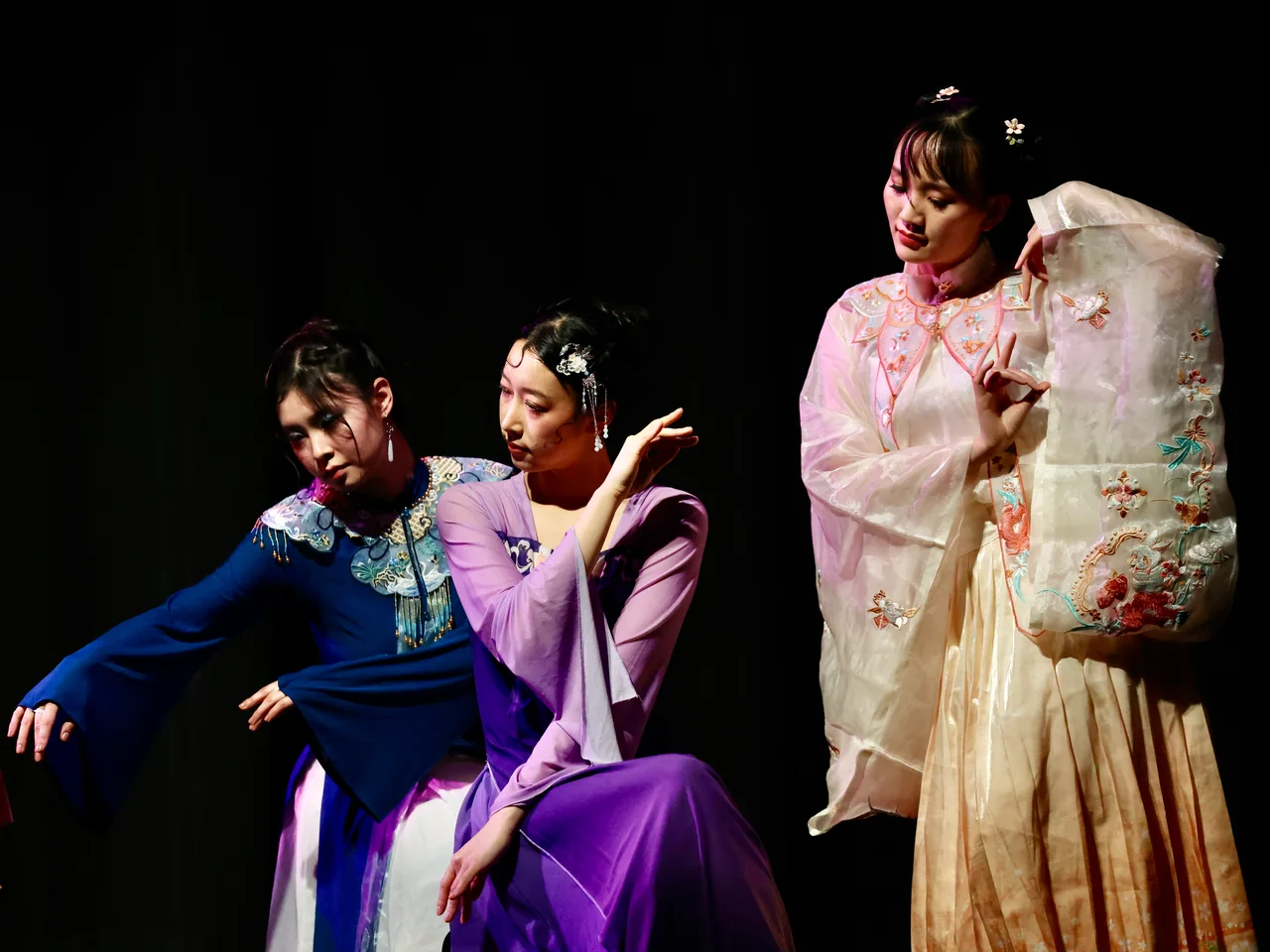 Three performers in blue, purple and cream leaning into a shared stage gesture