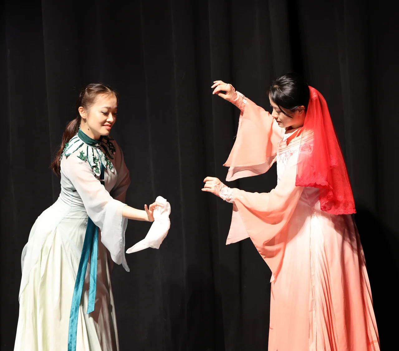 Two performers in pale green and peach reaching toward a white handkerchief
