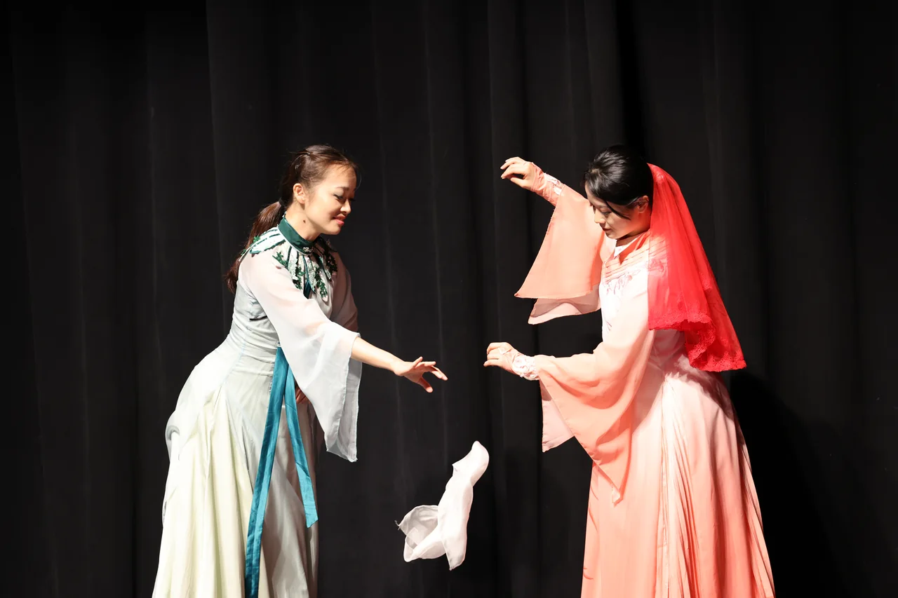 Two performers in green and peach costumes with a white handkerchief suspended between them
