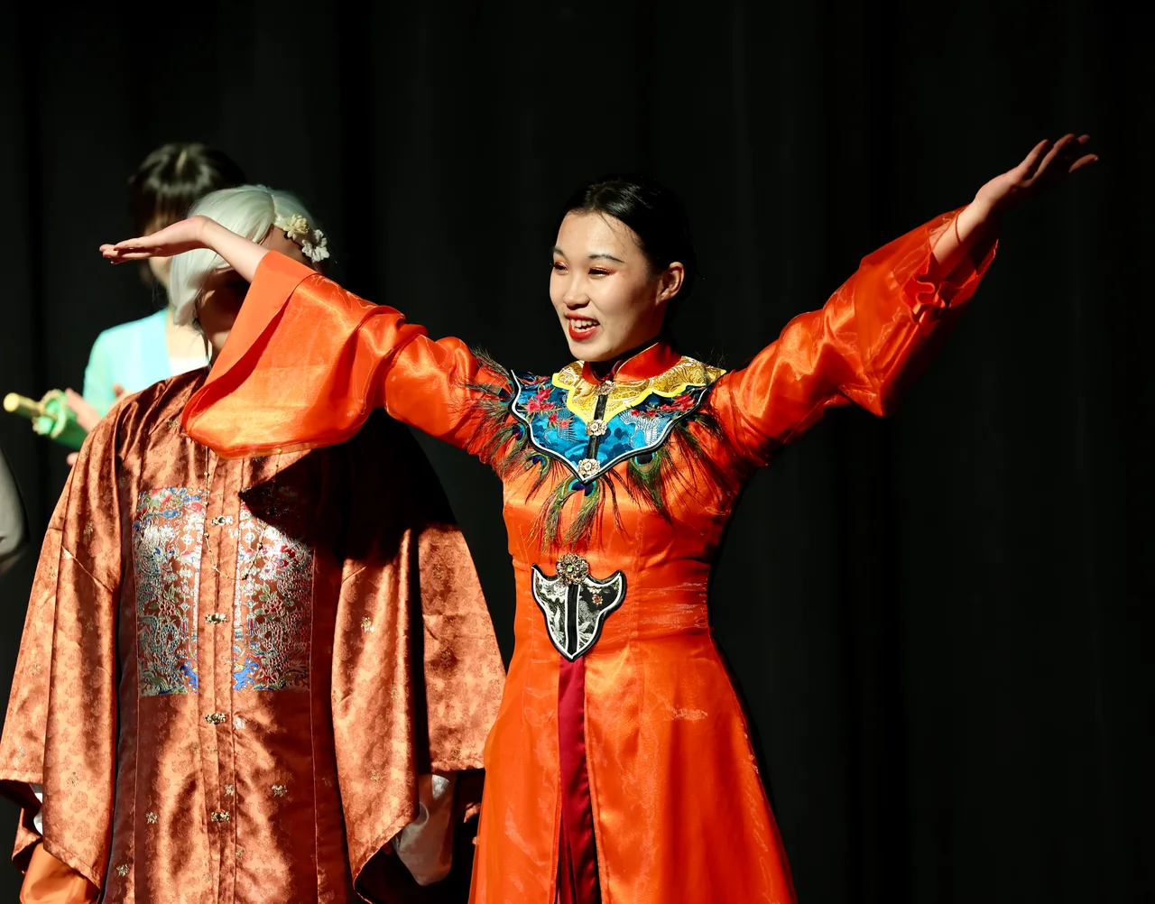 A performer in an orange embroidered costume smiling with arms spread wide